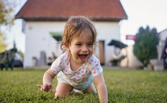 Picture of a happy baby crawling in the grass.  The baby is smiling and is shown with a house in the background. 