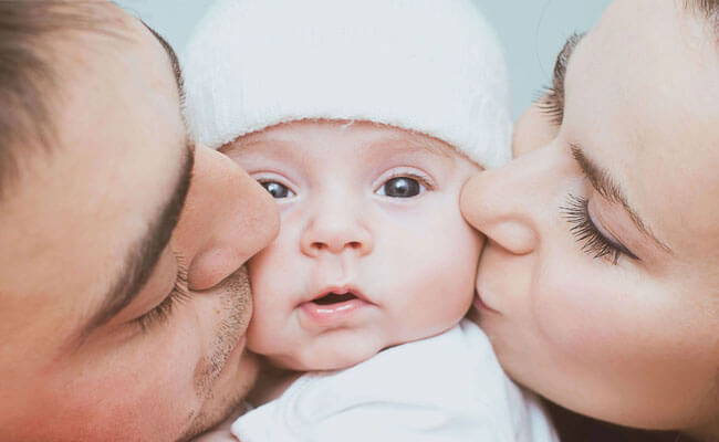 Picture of a happy couple kissing their baby. They are kissing the cheeks of the baby, who is looking directly at the camera.