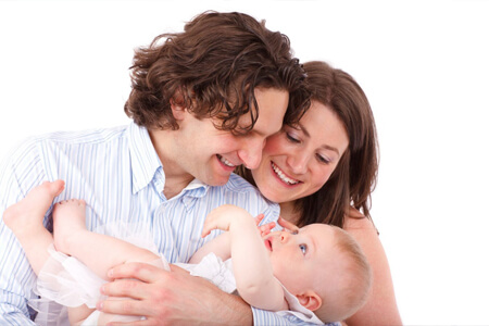 Picture of a young couple looking lovingly at their baby.  The man is holding the baby and both parents are smiling and looking at the baby.